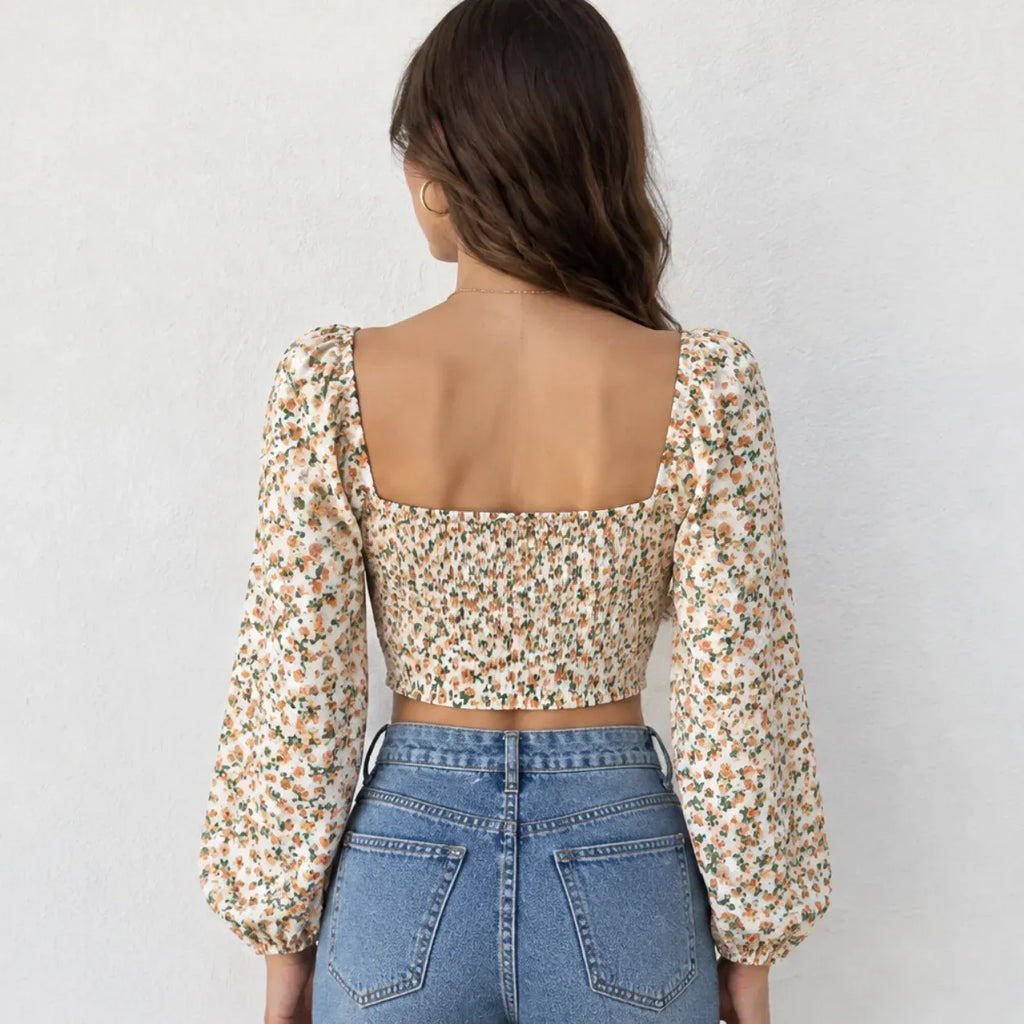 A woman shown from the back wearing a cream-colored puff-sleeve crop top with a dense orange and green floral print, paired with blue jeans, against a light gray textured wall.