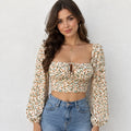 A woman wearing a long-sleeve cropped blouse with a small floral print in beige, orange, and green, paired with blue jeans, standing against a light gray textured wall.
