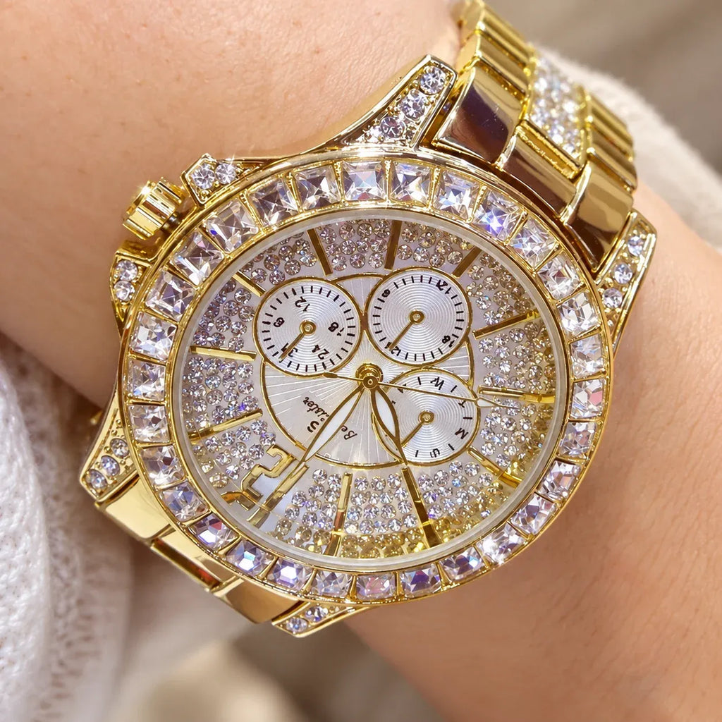 A gold-tone wristwatch with a bezel and link bracelet covered in clear rhinestones, worn on a wrist. The round white dial has three subdials and gold-tone hands and markers.