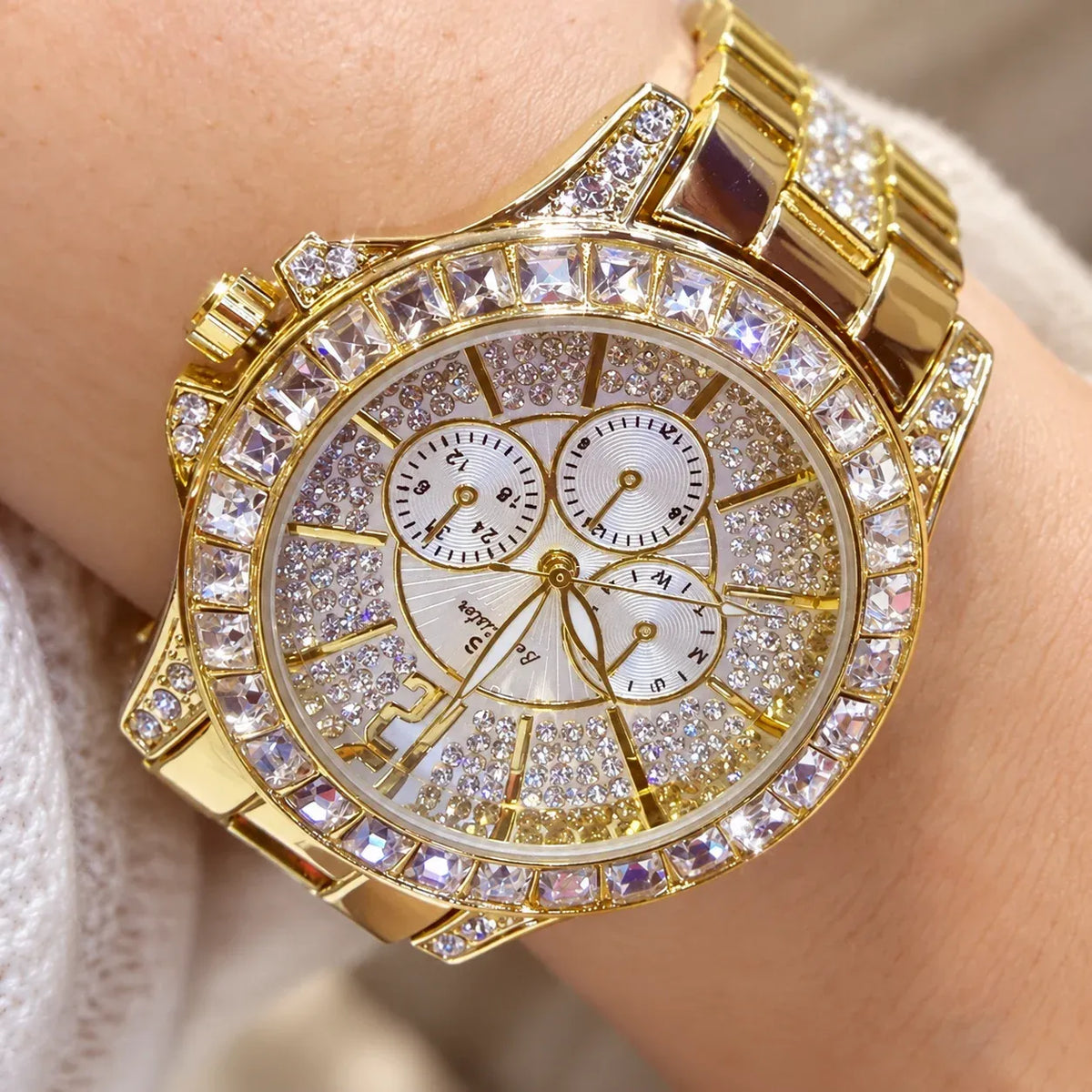 A gold-tone wristwatch with a bezel and link bracelet covered in clear rhinestones, worn on a wrist. The round white dial has three subdials and gold-tone hands and markers.