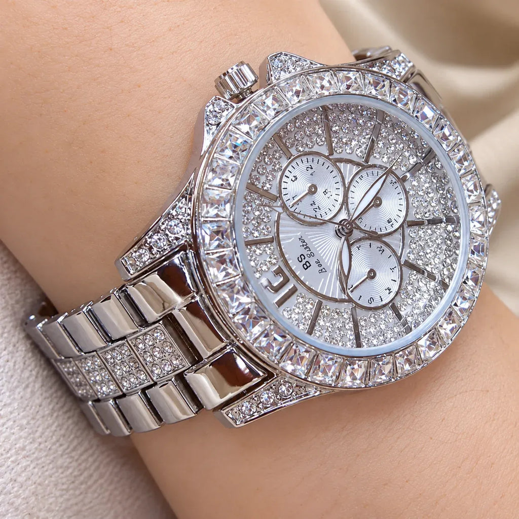 A close-up of a wristwatch with a silver-tone metal bracelet and case, heavily embellished with clear rhinestones. The round silver dial features three subdials and silver-tone hands.