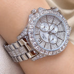 A close-up of a wristwatch with a silver-tone metal bracelet and case, heavily embellished with clear rhinestones. The round silver dial features three subdials and silver-tone hands.