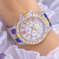 A silver wristwatch with a bezel and link bracelet covered in clear rhinestones, worn on a wrist. The round white dial has three subdials and gold-tone hands and markers.