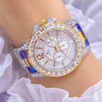 A silver wristwatch with a bezel and link bracelet covered in clear rhinestones, worn on a wrist. The round white dial has three subdials and gold-tone hands and markers.
