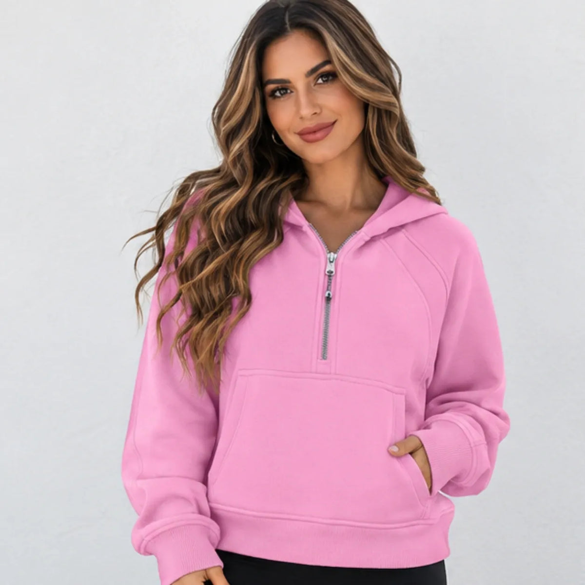 A woman wearing a pink half-zip hoodie with a silver zipper, kangaroo pocket, and ribbed cuffs, photographed against a light gray studio background.