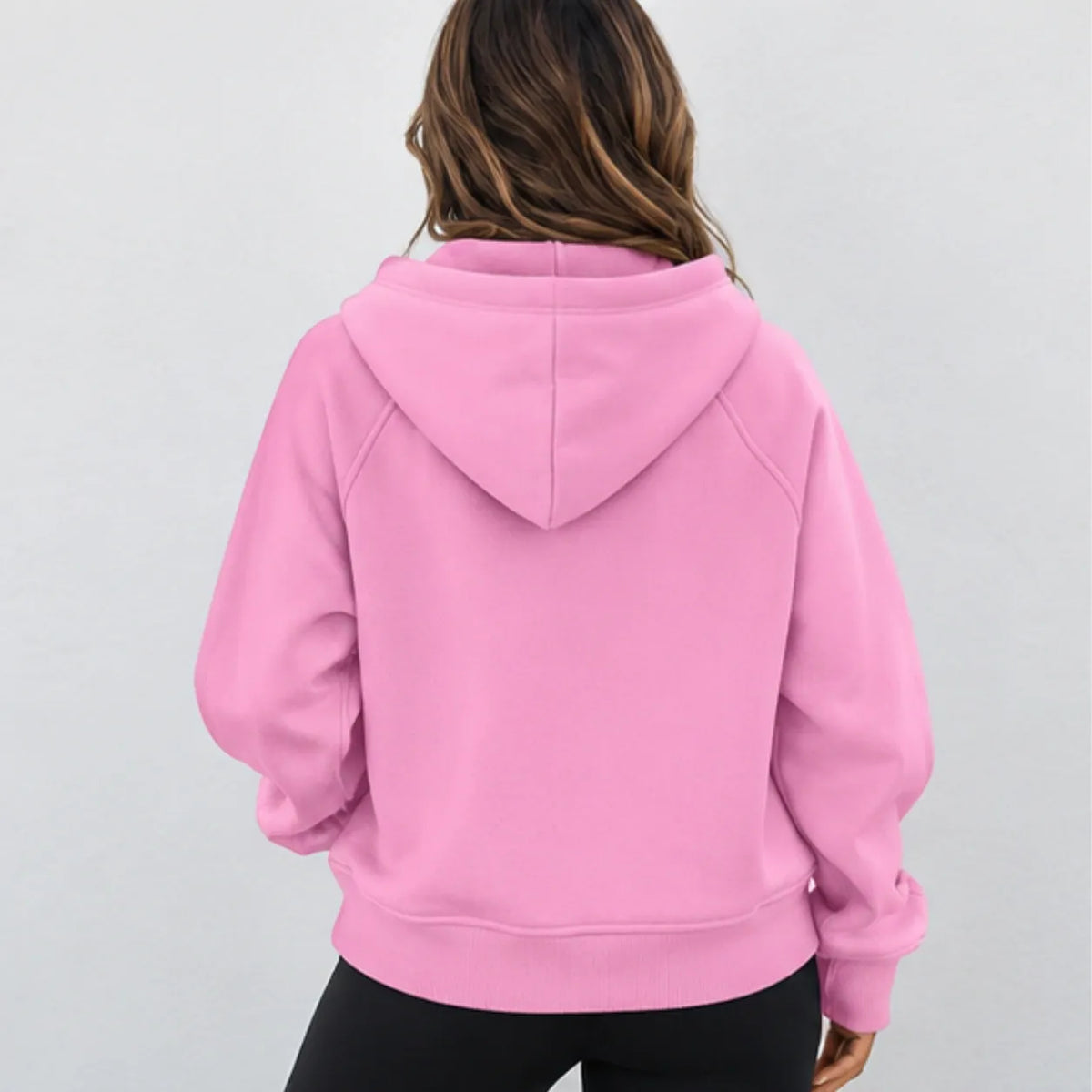 A woman viewed from the back wearing a light pink pullover hoodie with black pants, set against a plain light gray background.