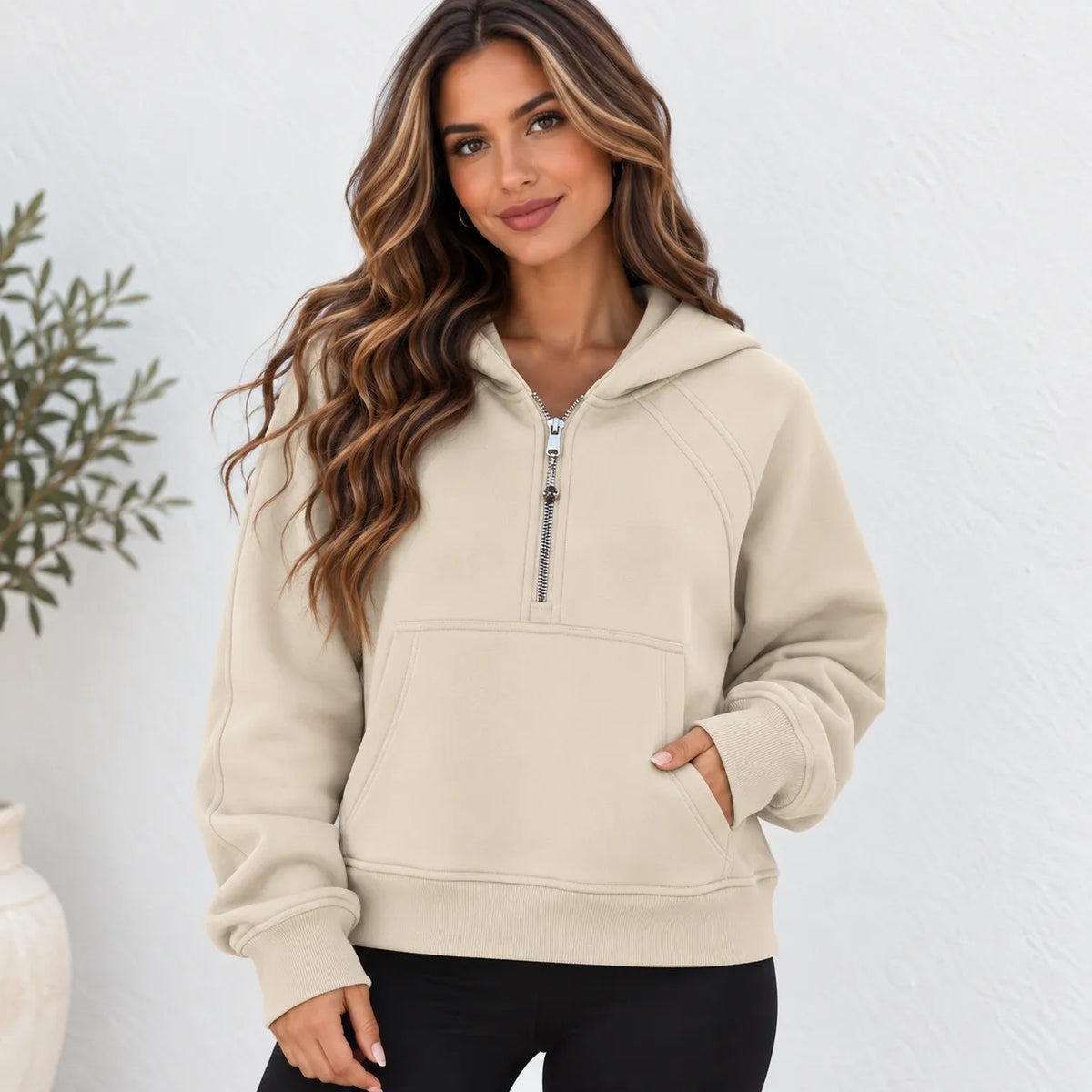 A woman wearing a beige pullover hoodie with a half-zip neckline and a front kangaroo pocket, paired with black pants, standing against a textured white wall.