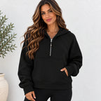 A woman wearing a black half-zip pullover hoodie with a silver zipper, kangaroo pocket, and ribbed cuffs, standing against a white textured wall.