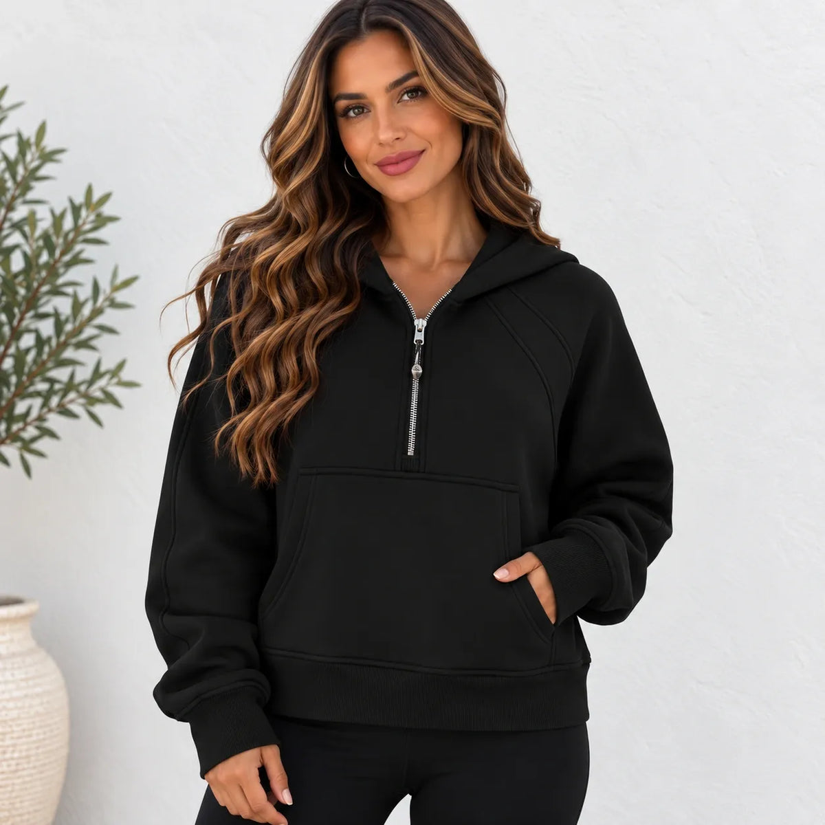 A woman wearing a black half-zip pullover hoodie with a silver zipper, kangaroo pocket, and ribbed cuffs, standing against a white textured wall.