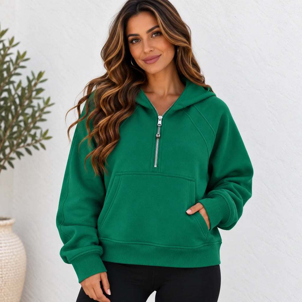 A woman wearing a bright green pullover hoodie with a half-zip front, kangaroo pocket, and ribbed cuffs and hem, paired with black pants, standing against a white textured wall.