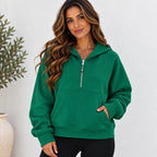 A woman wearing a bright green pullover hoodie with a half-zip front, kangaroo pocket, and ribbed cuffs and hem, paired with black pants, standing against a white textured wall.
