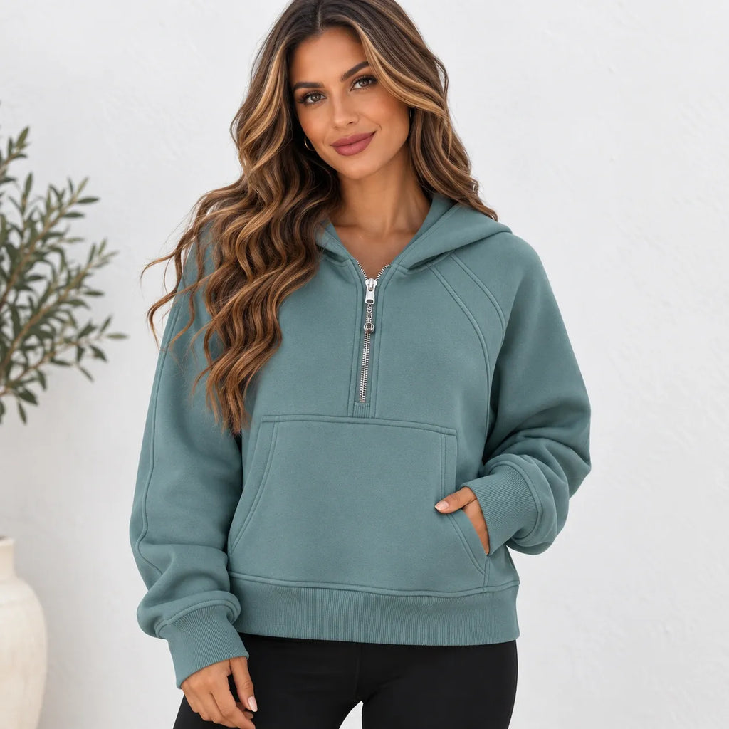 A woman wearing a sage green half-zip hoodie with a kangaroo pocket, standing against a white wall with a plant in the background.