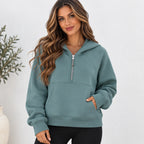 A woman wearing a sage green half-zip hoodie with a kangaroo pocket, standing against a white wall with a plant in the background.