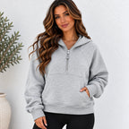 A woman wearing a light gray heathered half-zip hoodie with a kangaroo pocket, paired with black pants, standing against a textured white wall.