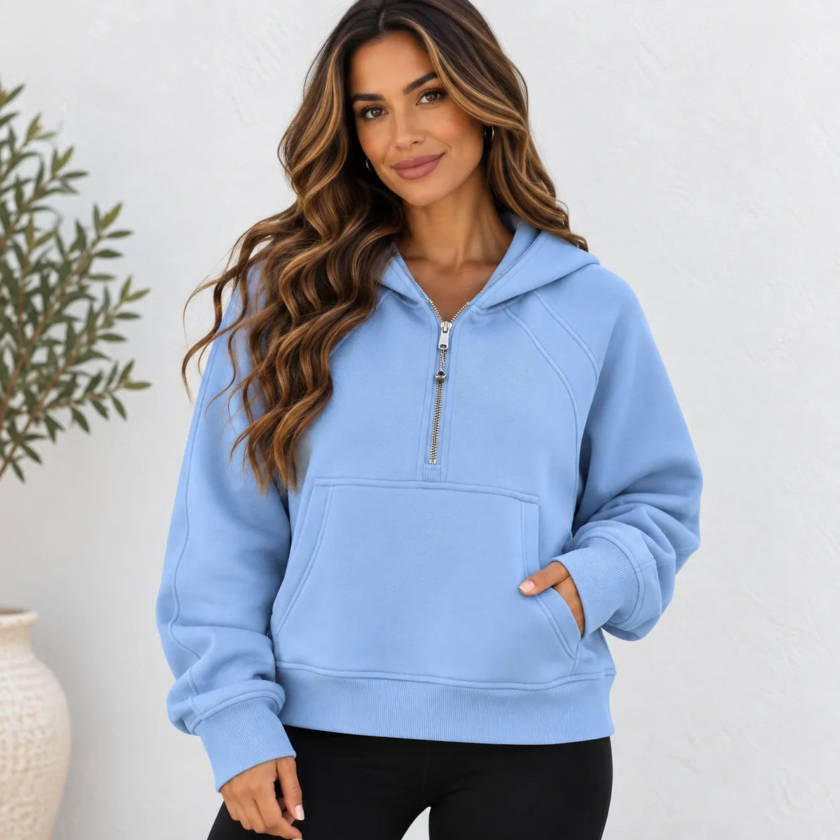 A woman wearing a light blue half-zip pullover hoodie with a kangaroo pocket, paired with black pants, standing against a white textured wall.