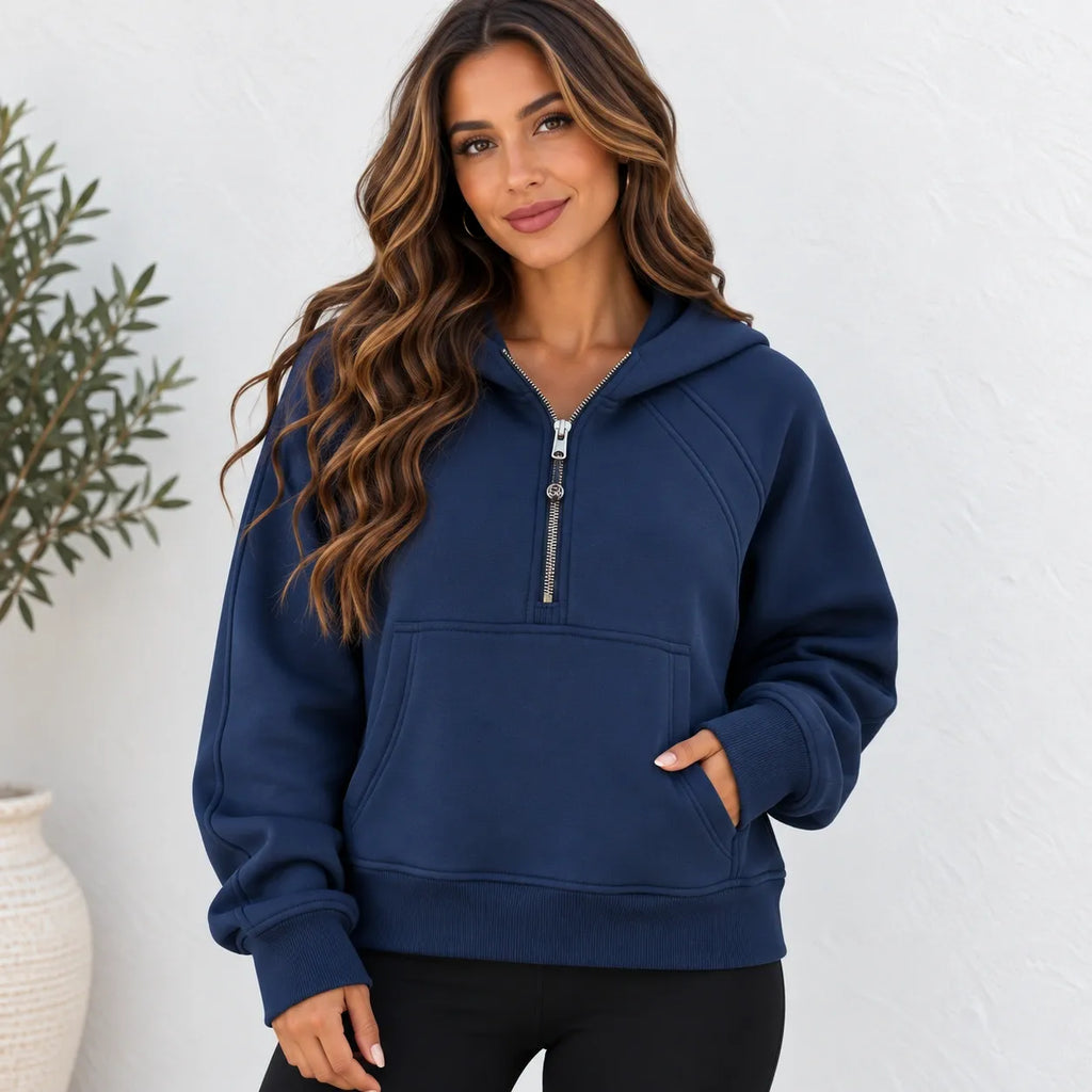 A woman wearing a navy blue half-zip hoodie with a silver zipper, standing against a white textured wall with a potted plant in the background.
