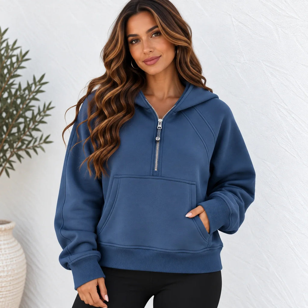 A woman wearing a slate blue half-zip hoodie with a front kangaroo pocket, standing against a white textured wall.