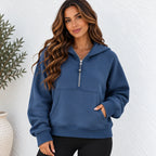 A woman wearing a slate blue half-zip hoodie with a front kangaroo pocket, standing against a white textured wall.