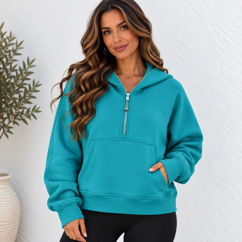 A woman wearing a teal turquoise half-zip hoodie sweatshirt with black pants, standing against a white textured wall with a potted plant on the left.