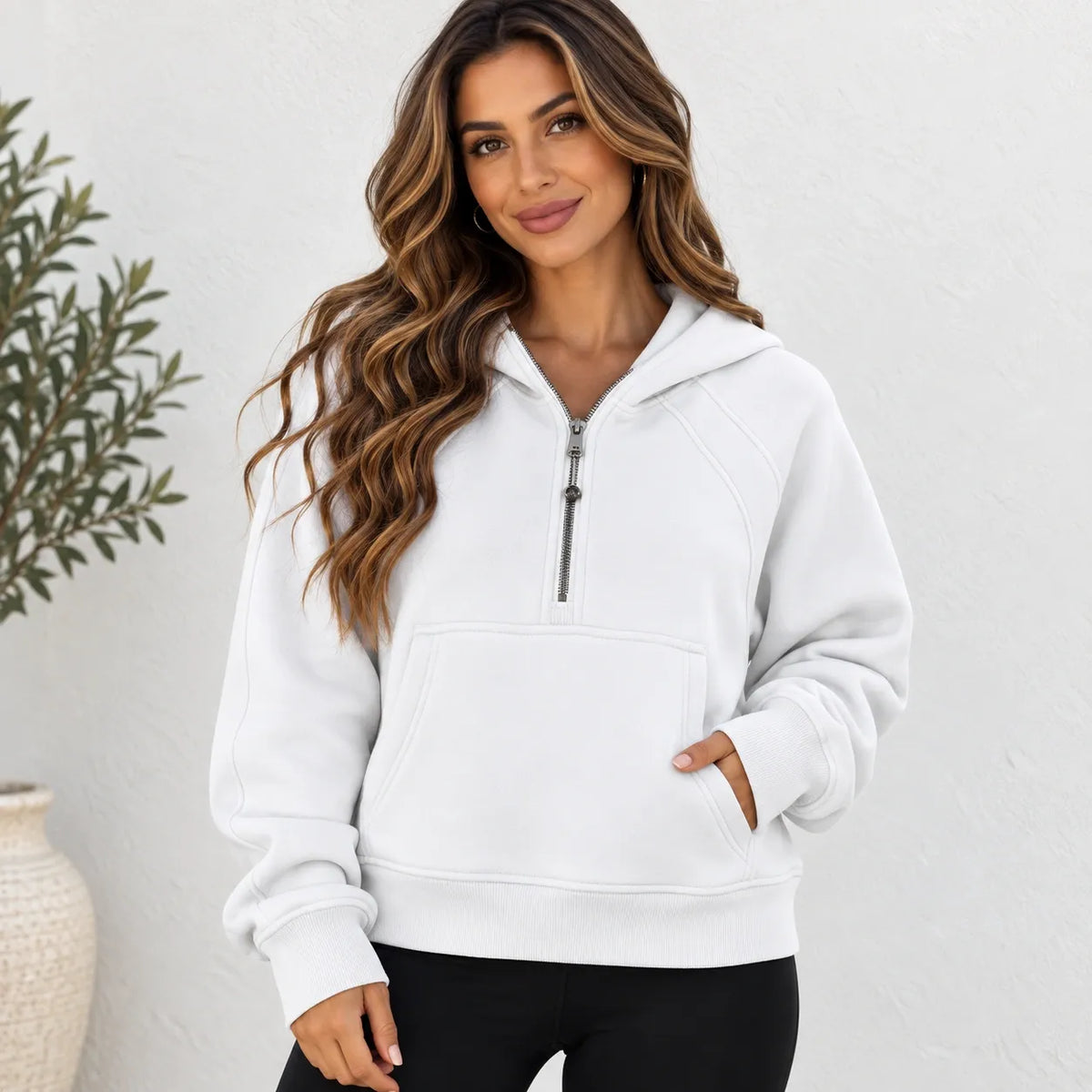A woman wearing a white half-zip pullover hoodie with a kangaroo pocket, paired with black pants, standing against a textured white wall with a potted plant on the left.