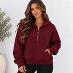 A woman wearing a burgundy half-zip hoodie sweatshirt with a silver zipper and a front kangaroo pocket, paired with black pants, standing against a white textured wall.
