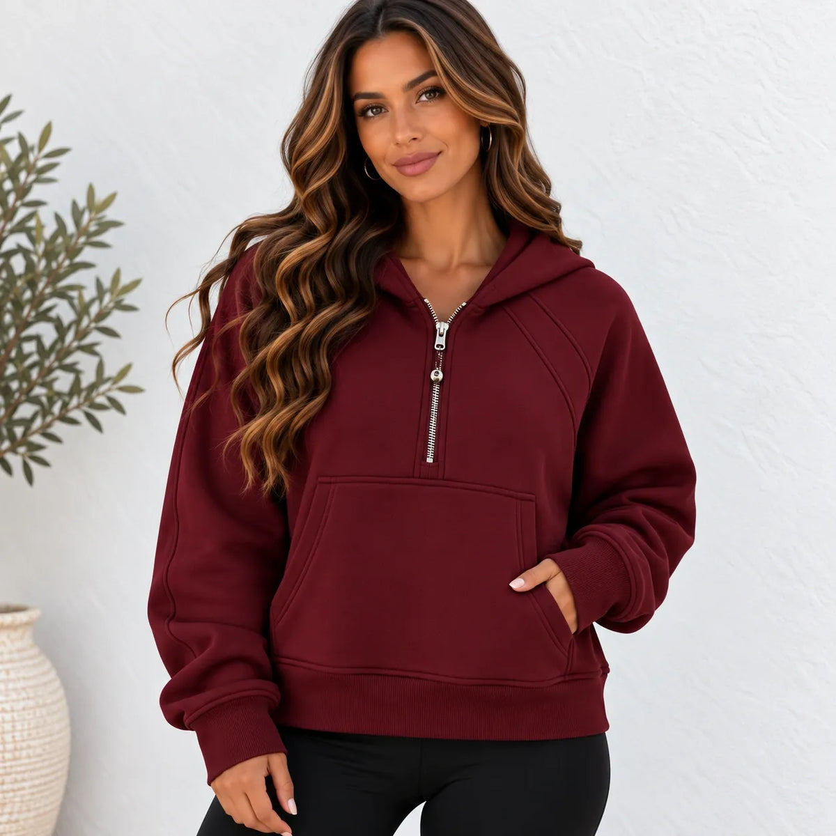 A woman wearing a burgundy half-zip hoodie sweatshirt with a silver zipper and a front kangaroo pocket, paired with black pants, standing against a white textured wall.