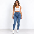 A woman modeling a pair of high-waisted, blue skinny jeans with a matching drawstring waist, paired with a white crop top and white sneakers, standing against a plain white wall.