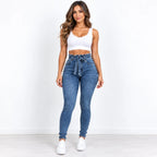 A woman modeling a pair of high-waisted, blue skinny jeans with a matching drawstring waist, paired with a white crop top and white sneakers, standing against a plain white wall.