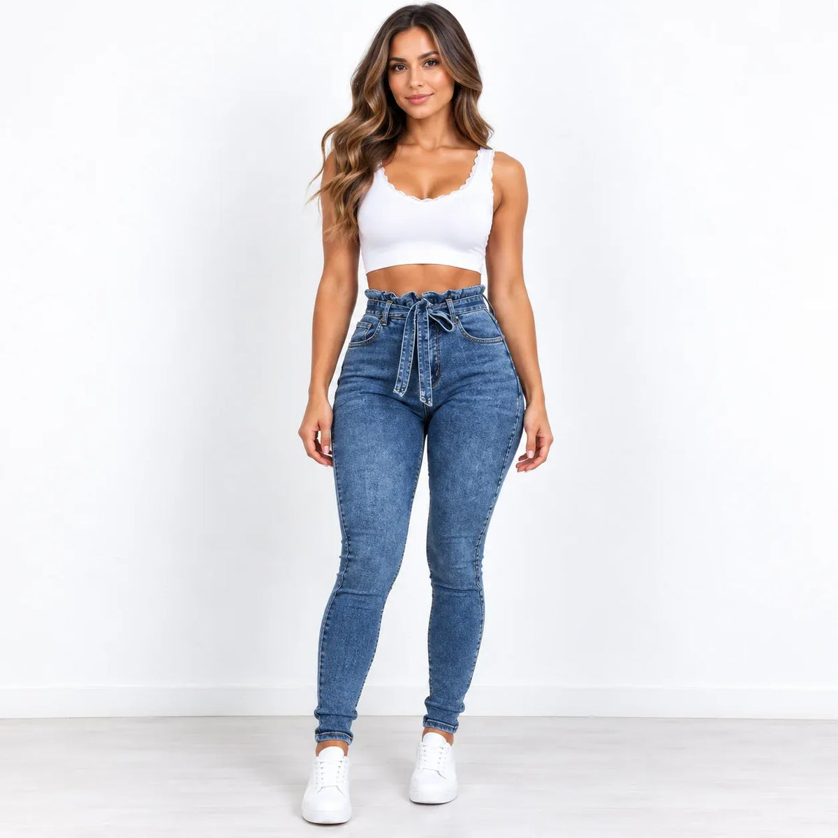 A woman modeling a pair of high-waisted, blue skinny jeans with a matching drawstring waist, paired with a white crop top and white sneakers, standing against a plain white wall.