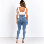 A woman is shown from behind wearing high-waisted blue skinny jeans, a white crop top, and beige high-heeled sandals against a white studio background.