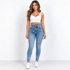 A woman modeling a pair of high-waisted, light-wash blue skinny jeans with white side stripes, paired with a white crop top and white sneakers, standing against a plain white studio background.