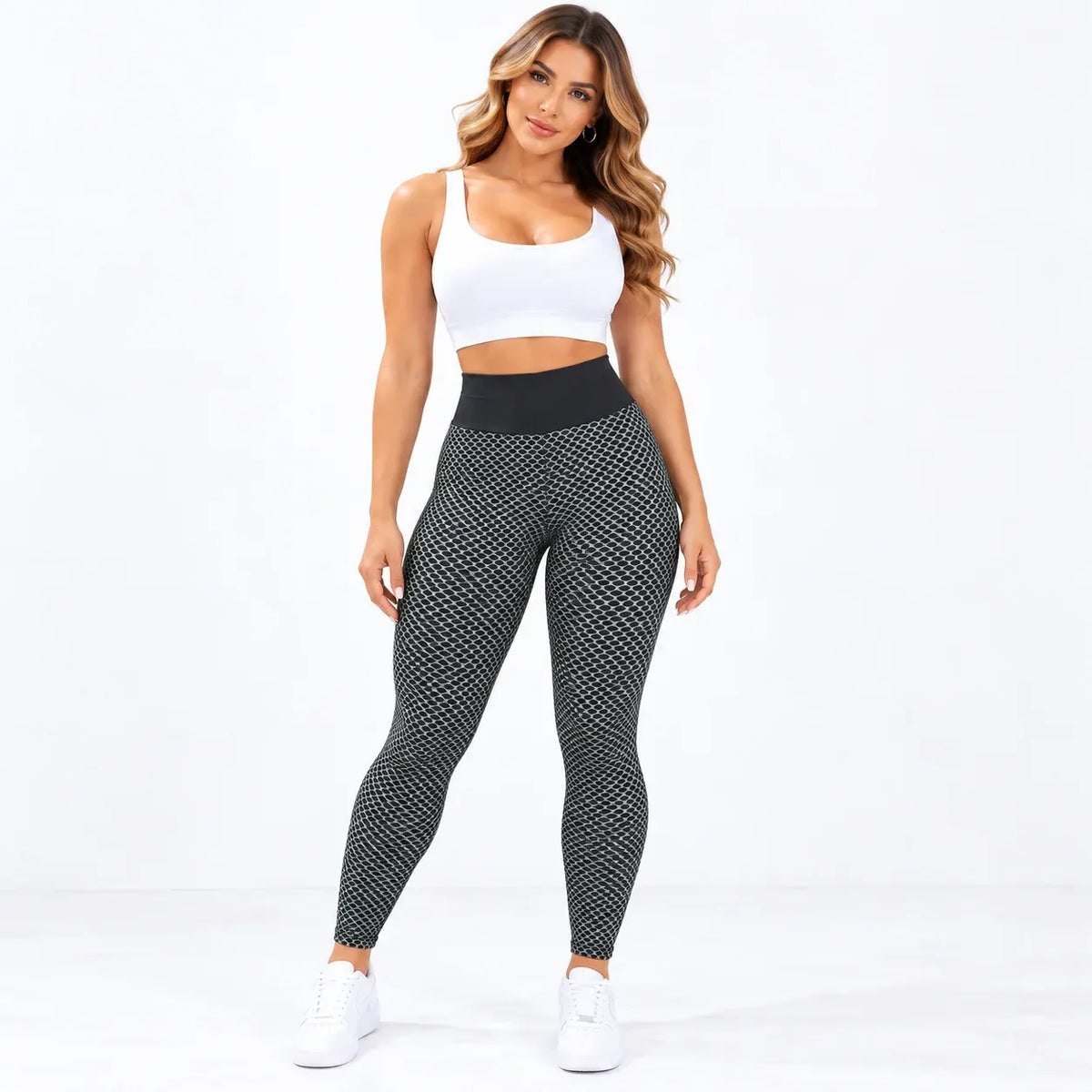 A woman modeling high-waisted black leggings with a white geometric pattern, paired with a white sports bra and white sneakers, against a plain white studio background.