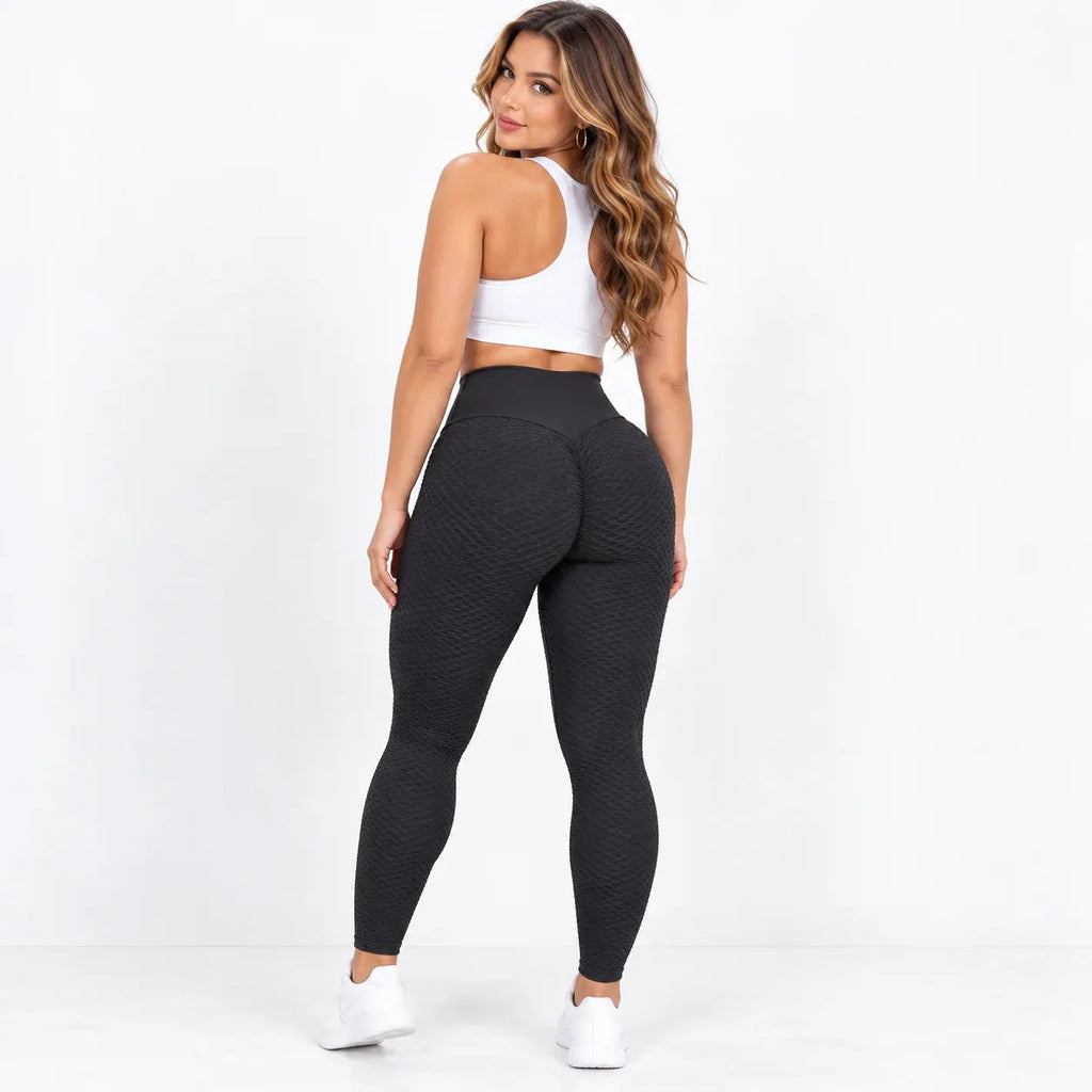A woman wearing textured black high-waisted leggings, a white sports bra, and white sneakers, photographed from behind against a clean white studio background.