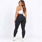 A woman wearing textured black high-waisted leggings, a white sports bra, and white sneakers, photographed from behind against a clean white studio background.