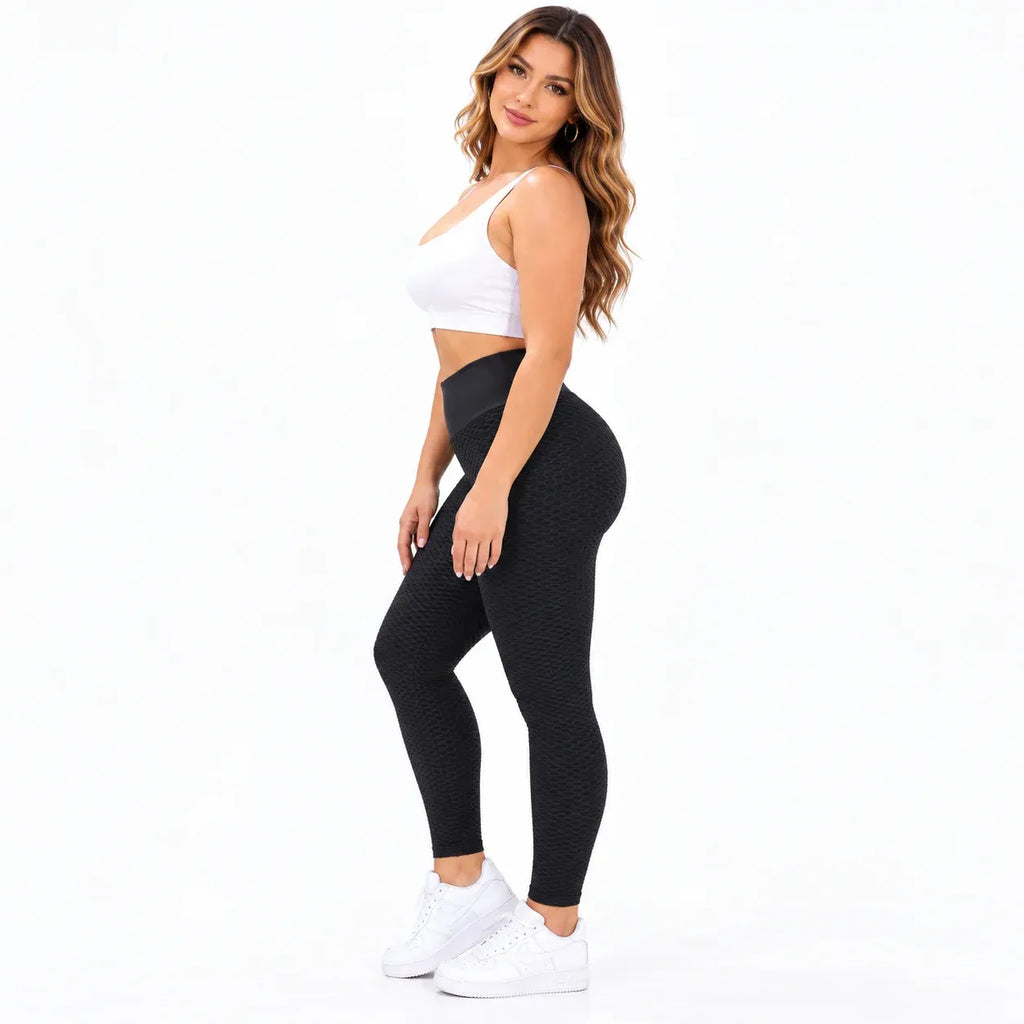 A woman modeling high-waisted black textured leggings, paired with a white crop top and white sneakers, standing against a solid white background.