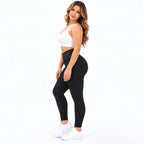 A woman modeling high-waisted black textured leggings, paired with a white crop top and white sneakers, standing against a solid white background.