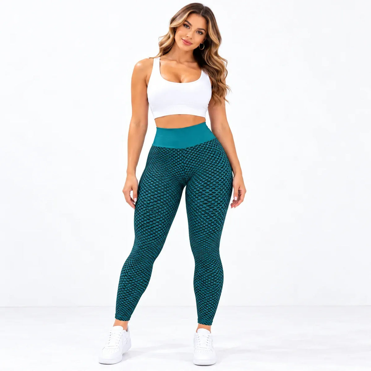 A woman modeling high-waisted teal patterned leggings with a solid teal waistband, paired with a white crop top and white sneakers, against a plain white studio background.
