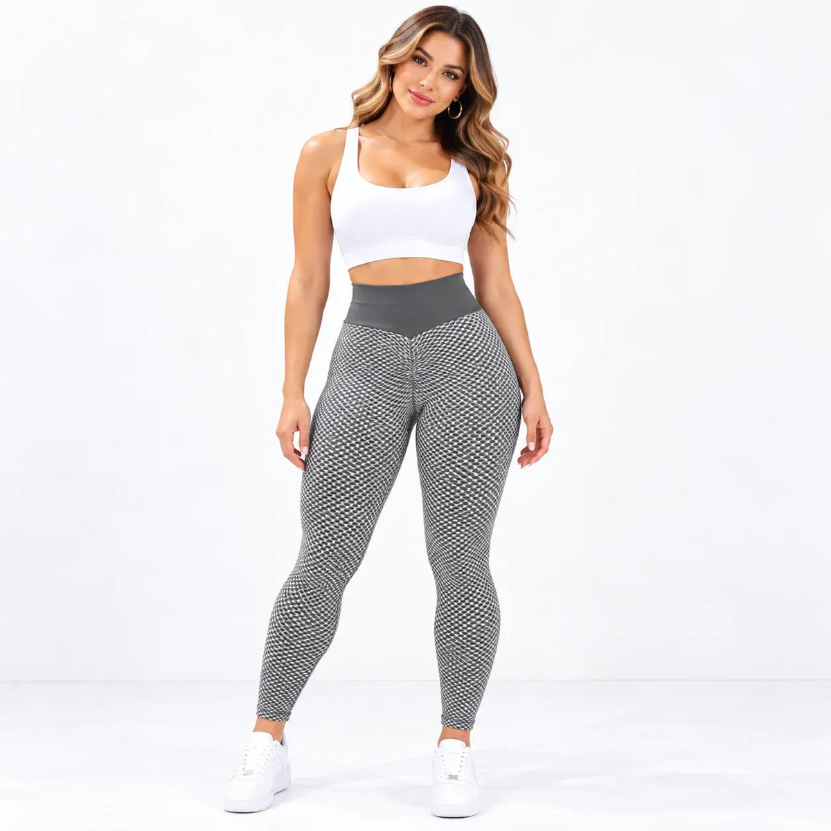 A woman modeling high-waisted athletic leggings with a gray and black geometric pattern, paired with a white sports bra and white sneakers, against a plain white studio background.