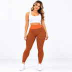 A woman modeling a two-piece activewear set consisting of a white scoop-neck crop top and high-waisted orange leggings with a black geometric pattern, paired with white sneakers against a white studio background.