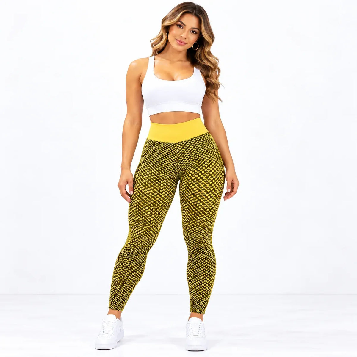 A woman modeling high-waisted athletic leggings with a yellow and black geometric pattern, paired with a white sports bra and white sneakers, against a plain white studio background.