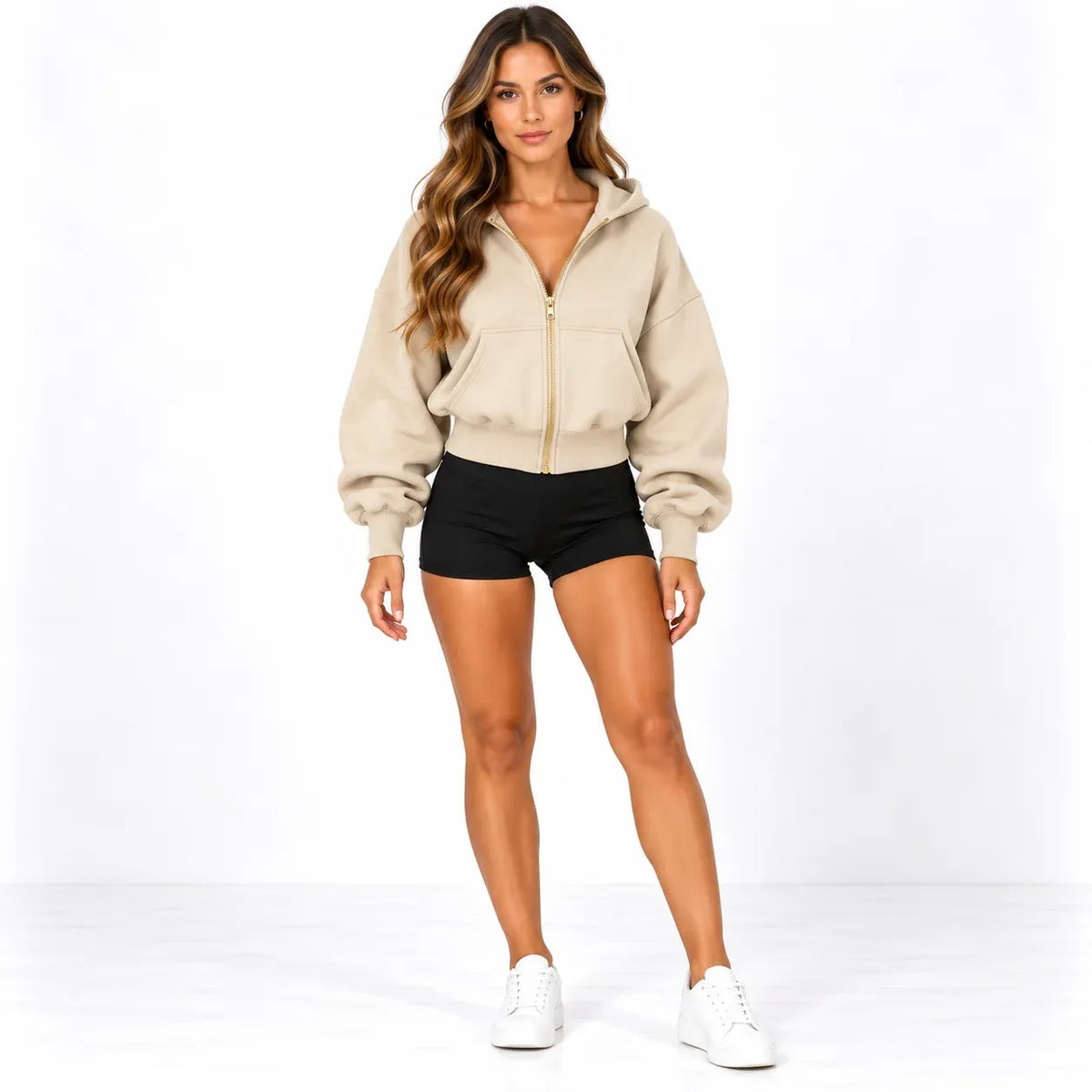 A woman modeling a beige cropped zip-up hoodie with a gold zipper, black athletic shorts, and white sneakers against a plain white studio background.