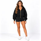 A woman modeling a black zip-up hoodie with a gold zipper and matching black shorts, paired with white sneakers, against a plain white studio background.