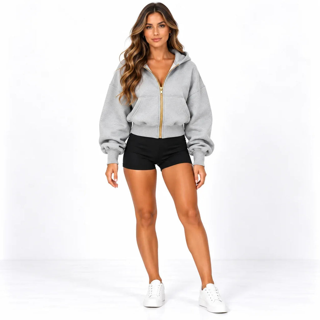 A woman modeling a light gray zip-up hoodie with a prominent gold zipper, paired with black shorts and white sneakers, against a plain white studio background.