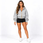 A woman modeling a light gray zip-up hoodie with a prominent gold zipper, paired with black shorts and white sneakers, against a plain white studio background.