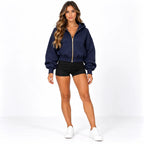 A woman modeling a navy blue cropped zip-up hoodie with gold trim, paired with black athletic shorts and white sneakers, against a plain white studio background.