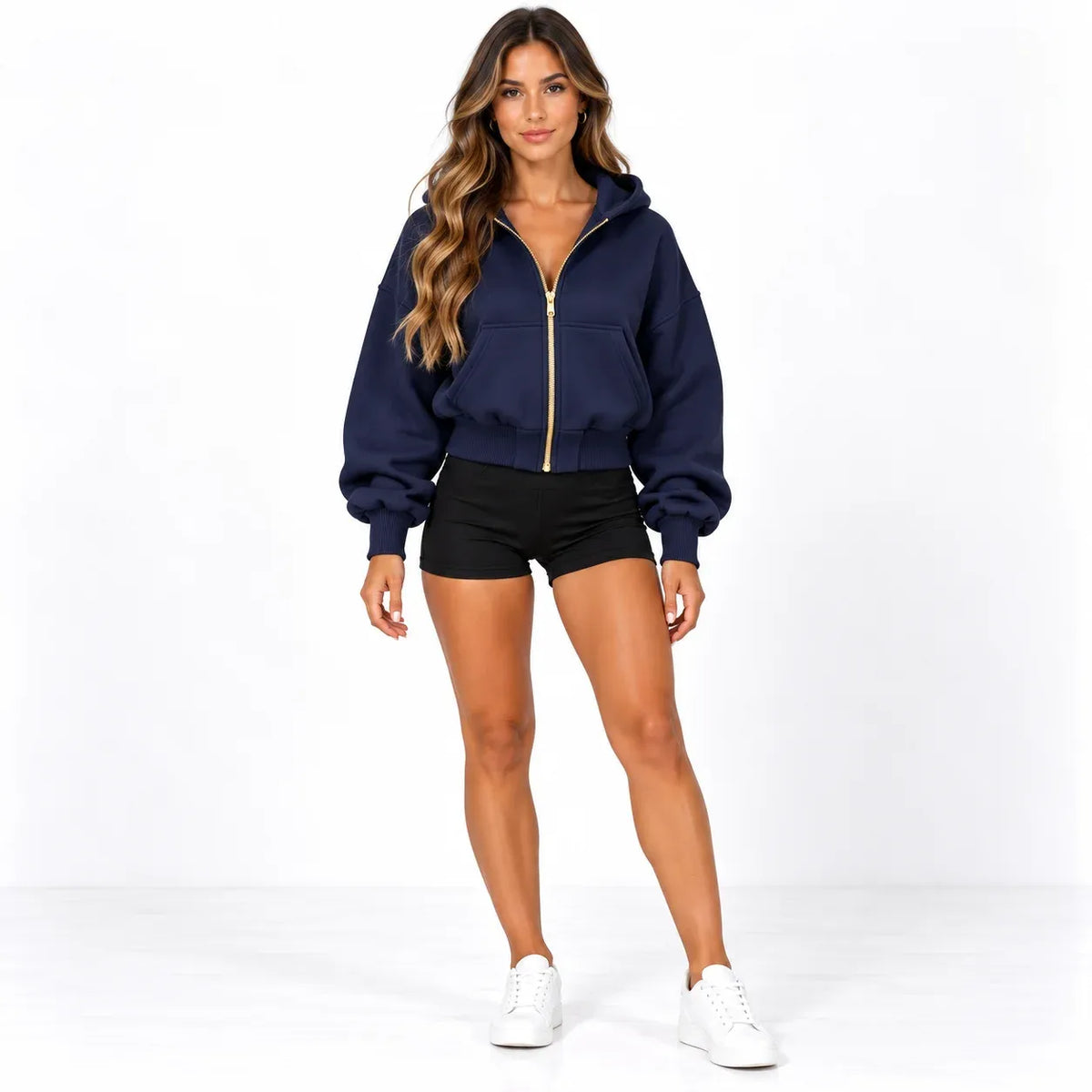 A woman modeling a navy blue cropped zip-up hoodie with gold trim, paired with black athletic shorts and white sneakers, against a plain white studio background.