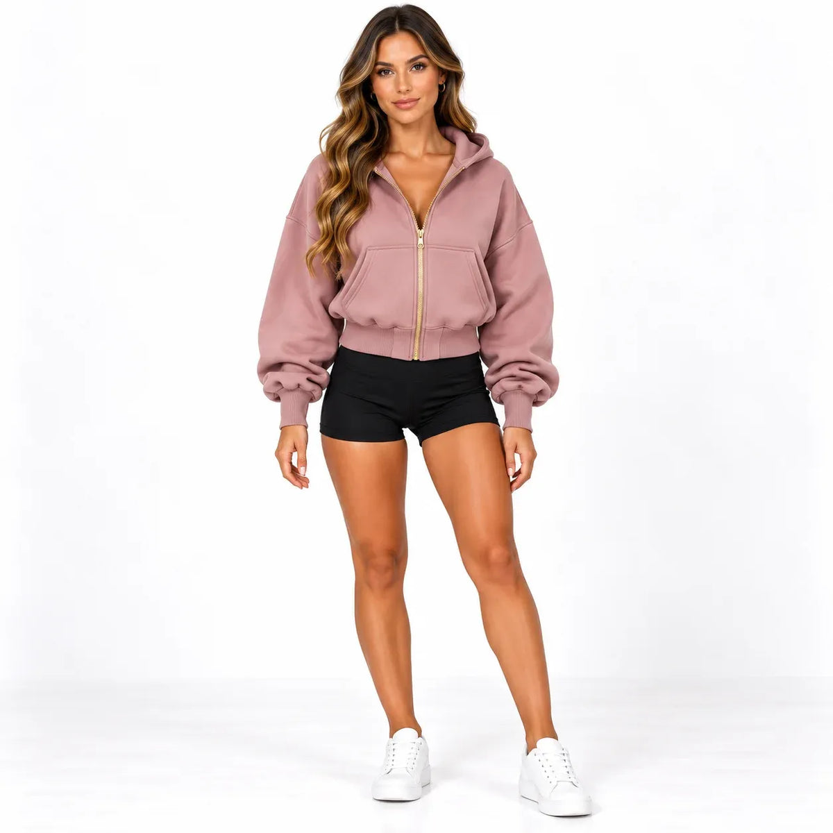 A woman modeling a dusty rose zip-up hoodie with a gold zipper, paired with black athletic shorts and white sneakers, against a plain white studio background.