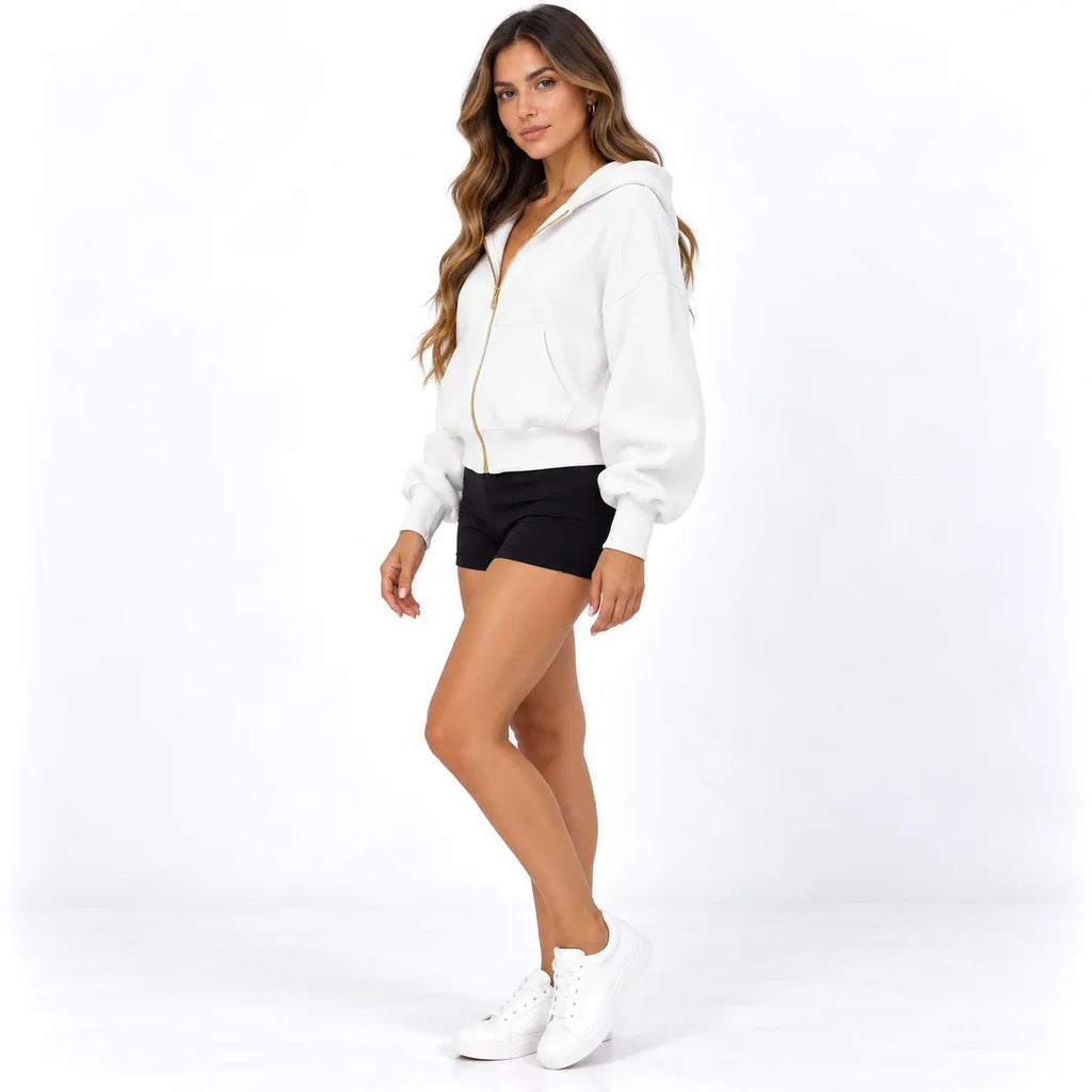A woman modeling a white zip-up hoodie with gold zippers, paired with black athletic shorts and white sneakers, set against a plain white studio background.