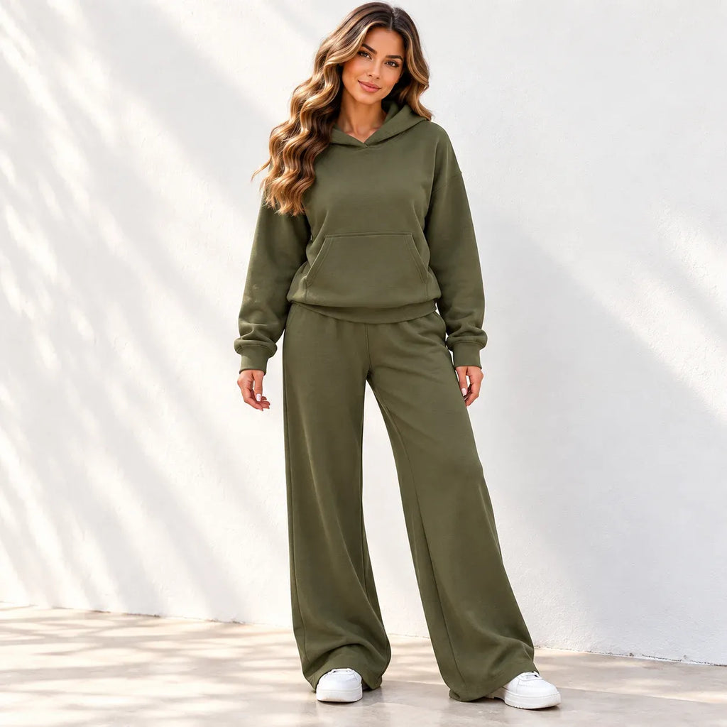 A woman modeling an olive green two-piece loungewear set consisting of a pullover hoodie and matching wide-leg sweatpants, paired with white sneakers against a white wall with soft leaf shadows.