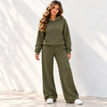 A woman modeling an olive green two-piece loungewear set consisting of a pullover hoodie and matching wide-leg sweatpants, paired with white sneakers against a white wall with soft leaf shadows.